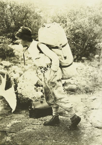 Carl carrying a large pack on his back and holding a bag. He is wearing a beaded belt that was almost certainly made by an Indian. 