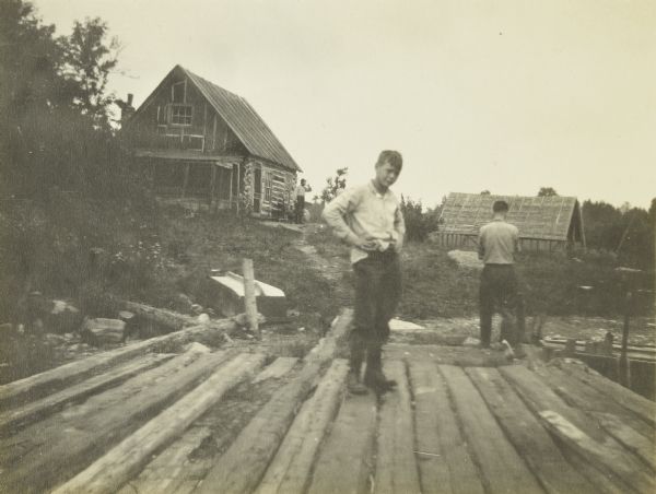 Jack standing on a pier with his hands on his hips. Behind him are two men and settlers' cabins.
