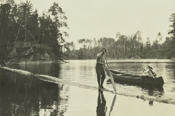 B (William Norris) walking a boom in the water with Jack looking on from a canoe nearby. B is using a paddle to balance himself. This is in the Narrows between Sand Point and Namakan Lakes.