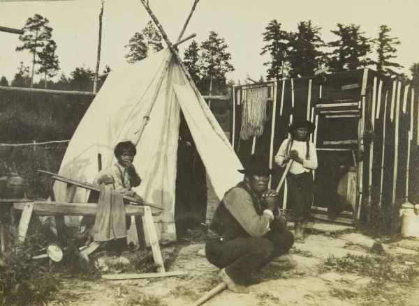 Two children and a man posing near a tipi (teepee) at an Indian settlement near Moose River.
