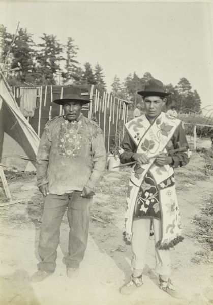 Two Indian men posing together at a settlement near Moose River. The man on the left is wearing an embroidered shirt that is probably made of leather. The man on the right is wearing beaded bandolier bags.