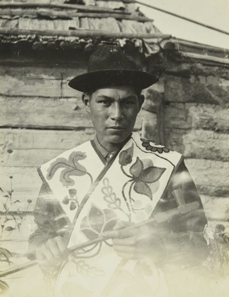 A young man wearing beaded bandolier bags. He is holding what appears to be a tobacco pipe.