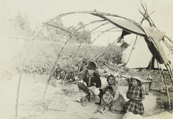 A father and his three young daughters posing together in an open structure made of branches.