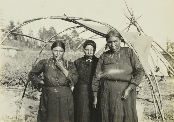 Three women posing together in a wickiup.