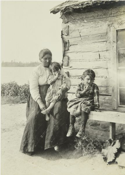 A woman posing with two young girls who are probably her daughters. They're sitting on a bench outside a log house, with the Moose River in the background.