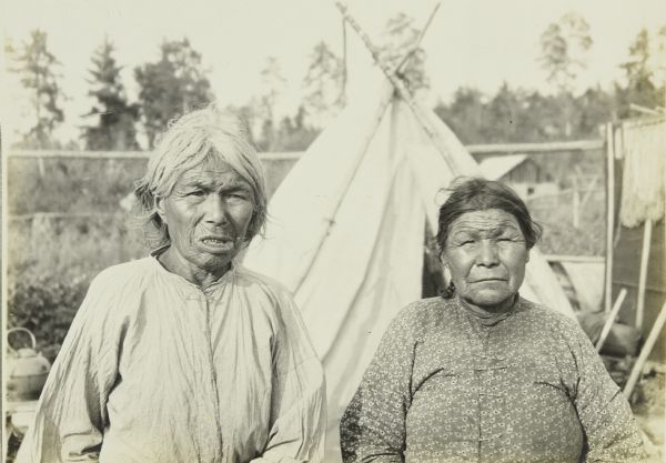 Two elderly women posing in front of a tipi (teepee) at a settlement near Moose River.