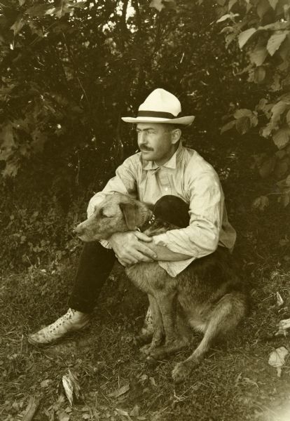 Bill Marr sitting on the ground outdoors with his arms around Carl's Airedale dog, Diadem (Di).