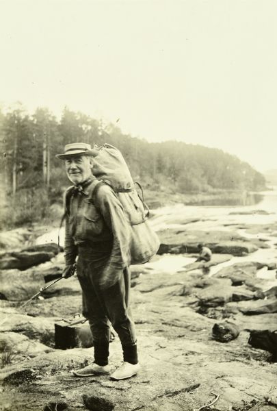 Billy Mac wearing a backpack while standing on rocks. He is holding a fishing pole, and another person is sitting on a rock near water in the background.