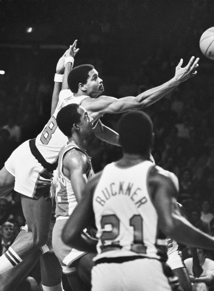 Marques Johnson of the Milwaukee Bucks sails past Dan Roundfield of the Atlanta Hawks and scores the bucket for two points. Quinn Buckner is watching the shot in the foreground.
