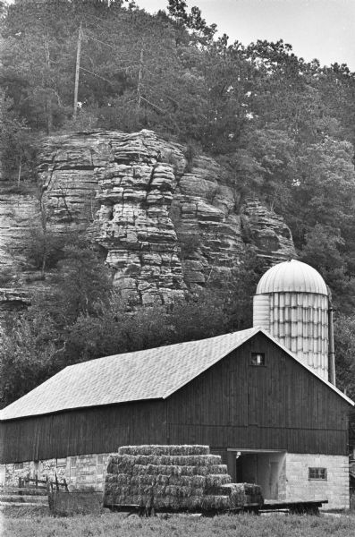 View across field towards a wagon stacked with bales of hay near a barn and a silo at the base of a bluff near Union Center.