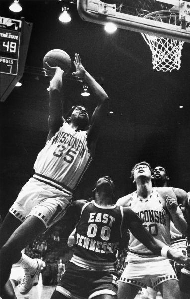 Claude Gregory showing good body control as he leaps off-balance for a layup over East Tennessee State guard Rusty Poole. Larry Petty is positioning himself under the basket for the rebound.