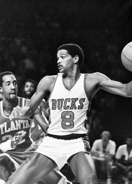 Marques Johnson of the Milwaukee handles the basketball while Dan Roundfield of the Atlanta Hawks guards him. In the background players are watching from the bench. 