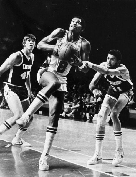 Marques Johnson of the Milwaukee Bucks splits two Denver Nuggets defenders and leaps for the layup as Bo Ellis (31) fouls him.