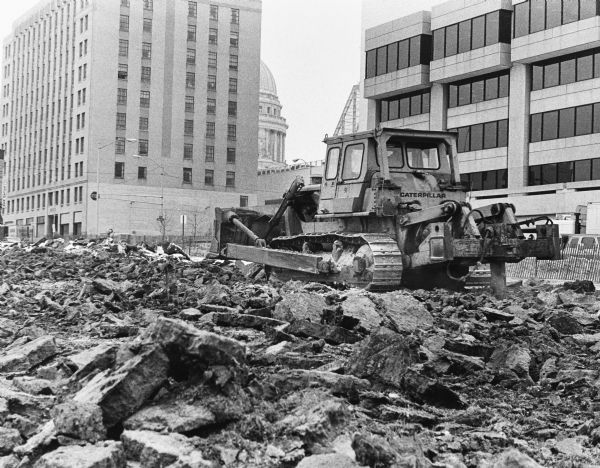 A Caterpillar bulldozer breaking ground for the construction of the State Natural Resources Building, also known as General Executive Facility (GEF) 2, and the State Education Building, or GEF 3, on South Webster Street. The Wisconsin State Capitol is in the background.