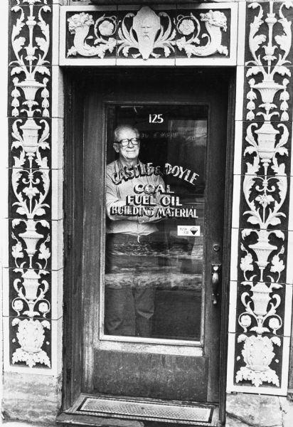 Jack Doyle standing inside the glass door of Castle and Doyle, Coal, Fuel Oil Building Material, on the 100 block of State Street in the historic firehouse building, with the original terra-cotta facade.