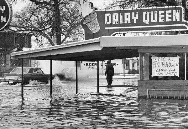 A man is walking through a flooded Dairy Queen parking lot in the downtown area, while cars are driving down the street. The sign in the window of the Dairy Queen reads: "Special Fishburger, catch your own, FREE." Across the street is a liquor store.