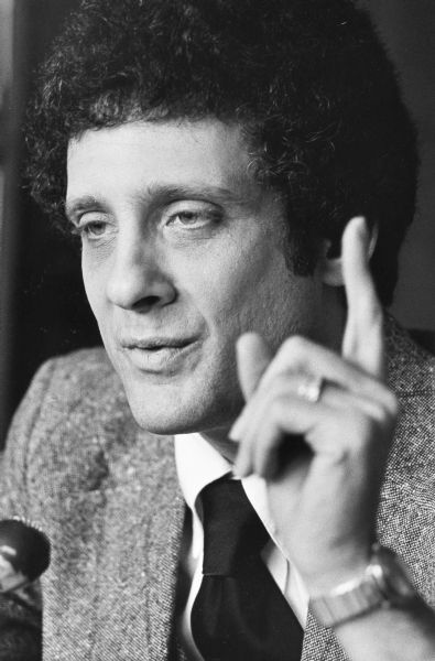 Journalist James Rowen speaking into a microphone and raising a finger during a talk.