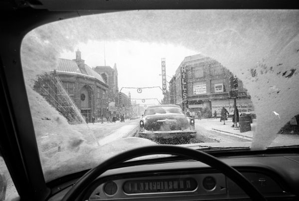 View of a snow storm through a windshield of an automobile while driving west down State Street. This was the first snow storm of the new year with 4 inches of snow expected. The Orpheum Theatre and Hill's Department Store are visible on the right and the Capitol Theater and Yost's Department Store are on the left.