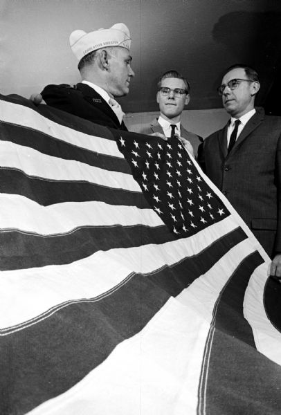 Charles A. Peckham from the Togstad Glen Disabled American Veterans chapter presenting an American flag to the Eastside English Lutheran Church Lutheran Pioneers, a chapter (train) of the national youth organization that has 108 chapters in the United States. In the center is David Marotz, train leader; and at right is Rev. Karl G. Bast, pastor of the church and train chaplain. The flag was given in memory of Henry Jopke and Robert Larson, two Madison men killed in service.