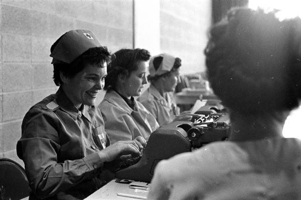 Three volunteer staff aides working at a Red Cross Bloodmobile. The women are dressed in Red Cross uniforms and caps and include, left to right: Mrs. James Hoag, Carolyn Durnford, and Mrs. John J. Stark. The women meet and register blood donors.