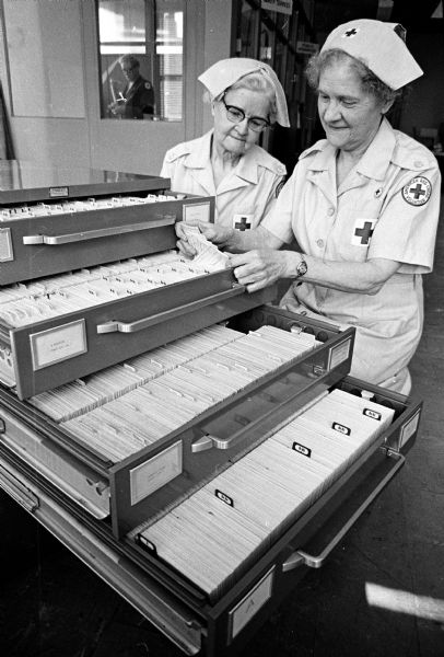 Two volunteers dressed in Red Cross uniforms and caps working with records of 22,000 blood donors. The women are Alma Wiese (left) and Violet Hughes.