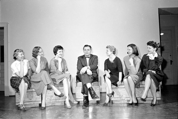 Six women are sitting with their legs crossed on either side of a man who is wearing a bow tie. The original caption states "King Henry VIII and his wives — as portrayed by Prof. Ray Stanley and members of Phi Beta alumnae — are shown before a rehearsal of 'The Royal Gambit', the next Phi Beta play reading." Left to right are Eileen Payne, Vera Sheppard, Mrs. Stuart Wirth, Prof. Stanley, Ruth Olson, Callie Stimson, and Lois Klein.