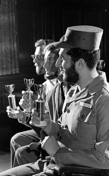 A profile view of three bearded engineering students who are posing and admiring trophies. The students, left to right, are John Menning, Dave Ludwig, and Faud F. Fuleihan. Fuleihan is wearing a Fidel Castro-type military cap.