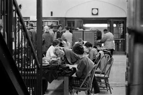 Young people crowded around library tables laden with stacks of books. The photograph is one of a series showing the crowded conditions at the Madison Public Library at 206 North Carroll Street, depicting the need for new library space.
