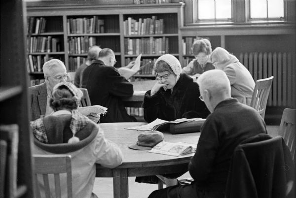 Men and women crowd around two library tables while reading. The photograph is one of a series showing the crowded conditions at the Madison Public Library at 206 North Carroll Street, depicting the need for new library space.