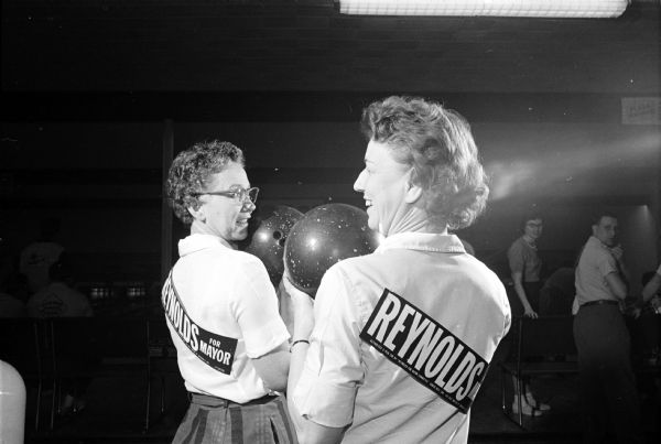 Two women holding up bowling balls pose in profile. The original caption states: "In a gesture of loyalty to their boss, Henry Reynolds, members of the Reynolds Transfer and Storage Co. bowling team have affixed campaign stickers to the backs of their team shirts." They are Min Martinelli and Lorraine Couture. Reynolds was running for mayor.