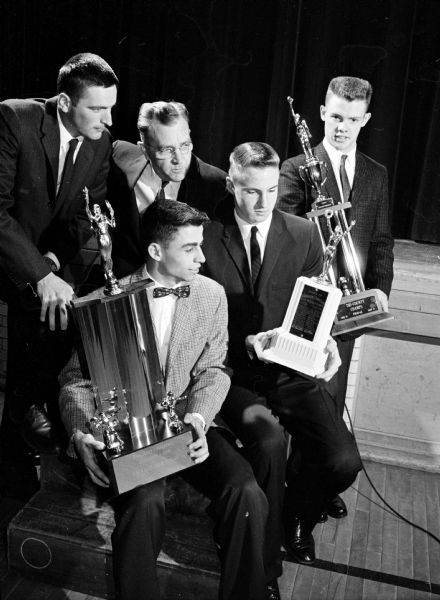Waunakee West High School athletes holding trophies. They include, left to right: Dick Barbian, football captain; Dave Roberts, most valuable basketball player; and John Howard, most valuable football player and basketball captain. In the rear, left, is Ron Hering, basketball coach and right, Larry (Moon) Mullins, Marquette athletic director and banquet speaker.