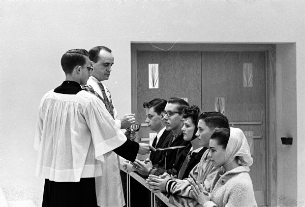 Five high school students participating in a anti-Communism crusade of young people from various parishes of the Catholic Diocese of Madison. The students attend mass every morning for a week at 6:30 AM. Students at the altar rail are, left to right, Mark Vitense, Ceaser Stravinski, Martha Reese, Allan Gurriell and Sandy Crimmins.  Rev. Leo Joyce is distributing the elements, assisted by Terry Welsh.