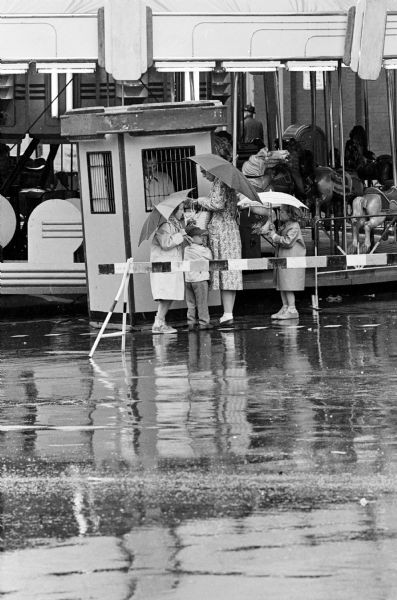 A woman and three children standing under umbrellas behind a barrier in the street during Madison's Outdoor Living Week activities.
