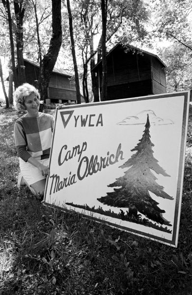 Peggy Moore is shown with the new sign which will mark the entrance to the YWCA's Camp Maria Olbrich on Lake Mendota.