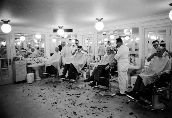 Four barbers dressed in white are cutting the hair of four men sitting in barber chairs. The barbershop, located in the basement of the State Capitol, was owned by Larry Brilliott. "The walls, floors, and ceilings of the shop are of marble, like the rest of the building. Most of the walls are covered with mirrors." Not shown, but mentioned in the text, is that "among the furnishings is a back room with showers and tubs."