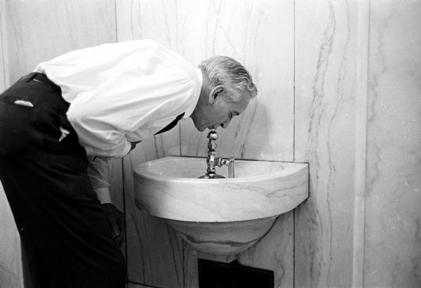 A man is bending over to drink from a metal water fixture. Water from the fixture spouted up and back onto the fixture, supposedly making it "sterile." These are known in Wisconsin as "water bubblers." The drinking fountain had a marble basin and was run by compressed air. The photo caption reads: "Almost as good as a cool martini."