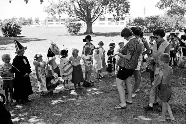 Children in costumes line up as three mothers judge their costumes. The finalist contestants are dressed as assorted characters including a hula dancer, an Indian maiden, tramps, and cowboys.