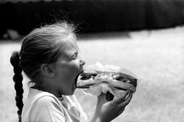 Rhonda Swenson, age 10, takes a bite of a king size hot dog at the Dane County Junior Fair. She is the daughter of Mr. and Mrs. Gerhard A. Swenson, 1318 E. Mifflin Street.