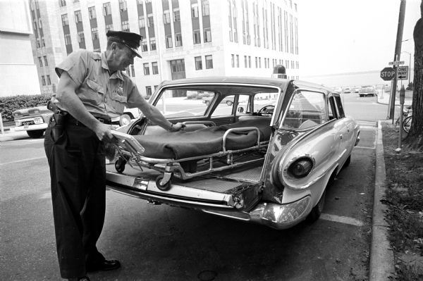 Officer Howard (Pete) Stormer displaying the interior of the new station-wagon sized Madison police ambulance. For the past 5 years the department relied on private ambulance services, but has now returned to having three of its own. Taken on S. Carroll Street at the intersection of W. Wilson Street. The building in the left background is the State Office Building with Lake Monona behind it.  