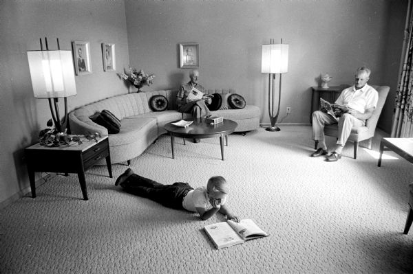 David Saalsaa, age 9, studies while his Uncle Oscar, left, and father, Edward relax and read in their shared living room. The room has a Mid Century curving sectional sofa and Mid Century floor lamps.
