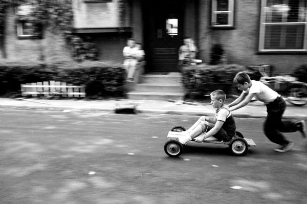 Peter Schram, 10, sitting on a home-made race car while being pushed by Bruce Mills, 11. The boys built the racer from boards and wheels from an old coaster wagon. They are testing their soapbox racer in the street on a dead end of South Brearly Street.