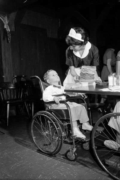 Present at an open house and party at the Fireside Pancake House, 625 State Street is Michael Aberle, 4. He is seated in a wheelchair and looking at waitress Terry McCormick who is carrying a plate piled high with pancakes. The open house is sponsored by the restaurant and United Cerebral Palsy of Dane County for children with cerebral palsy and their parents.