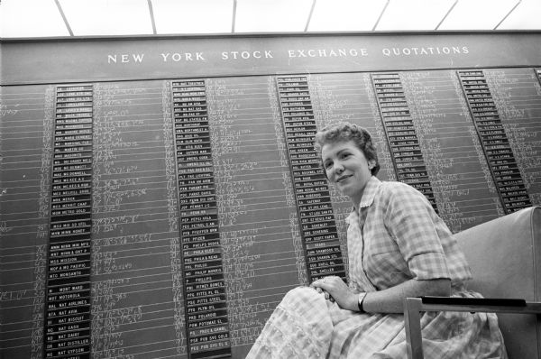 Mrs. William (Martha A.) Robbins, 4325 Wakefield Street, sitting at a broker's office in front of a New York Stock Exchange quotation board. Robbins is an active member of Madison's pioneer women's investment club made up chiefly of fellow employees at CUNA Mutual.