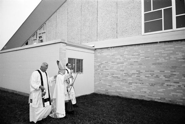Three Catholic clerics bless the exterior of the new St. Joseph's parish building at 1905 W. Beltline Highway. Bishop William O'Connor, wears robes and a miter (bishop's hat) while holding a staff in one hand and using a holy water sprinkling device with the other. He is assisted by two other priests also wearing robes. They are the Rev. Leonard Wagner, left, and the Rev. Nicholas Nirschl.