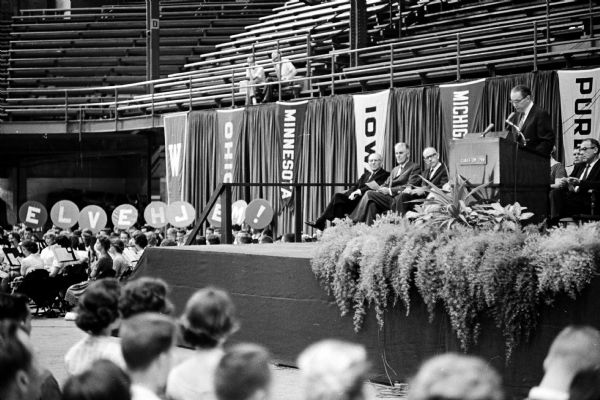 U.W. President Conrad Elvehjem speaks at the podium on the Field House stage. He advised the students to "buckle down" to their studies. On the left, tuba players have the letters spelling out the president's name on the covers of their tubas.