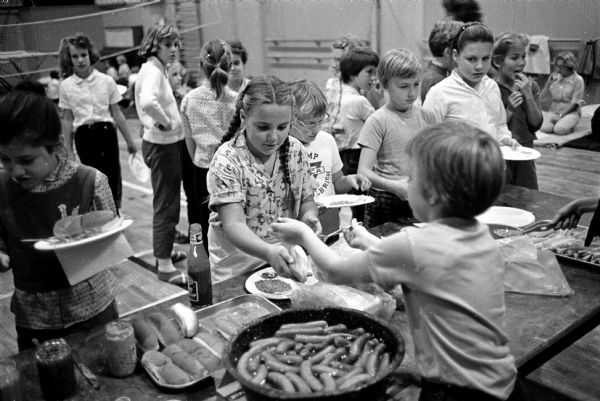 Cold weather sent a YWCA camping experience indoors for the 49 girls attending. Those in line for camp-type food of wieners and baked beans include Annette Wilcox (right) popping a hot dog into a bun for Martha Haygood (left).