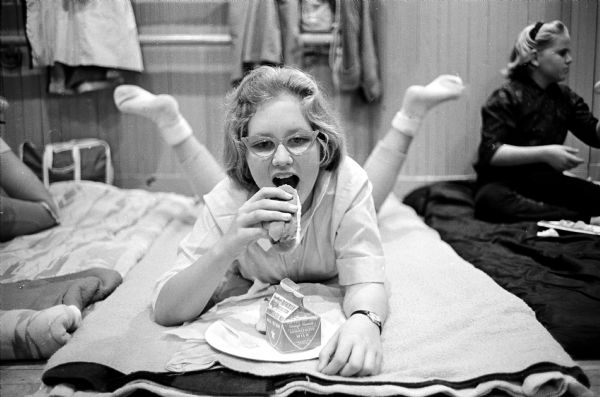 Jennifer Jueds is shown enjoying milk and a hot dog as she relaxes on her "bed" in the gymnasium of the YWCA. She is one of 49 girls who spent 24 hours camping indoors when the weather prevented their planned outdoor camping.