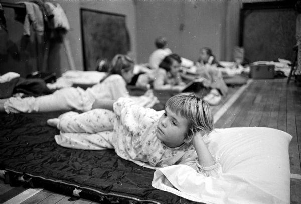 Patty McCutchin is shown attentively listening to a story while resting on a bedroll in the gym of the YWCA. Other pajama-clad girls are nearby reading or listening. Weather sent their camping experience indoors.