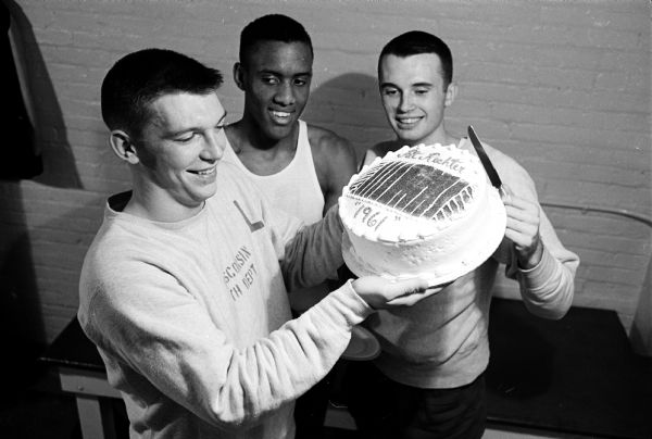 Members of U.W. basketball team presenting a cake to their new basketball teammate, Pat Richter, at a party in honor of his being named to the UPI All-American football team. Richter is shown holding the cake, decorated to show a football field with his name and "1961" in frosting. With him are Tom Gwyn (center) and Ken Siebel.