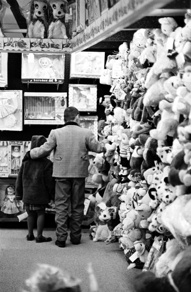 A man and child looking at a toy display in a store. On the wall next to them is a large display of puppets and stuffed toys. 
